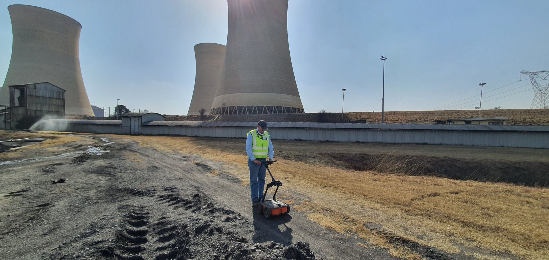 An HD GPR technician pushing a ground penetrating radar cart across the ground at an industrial site, with large cooling towers behind