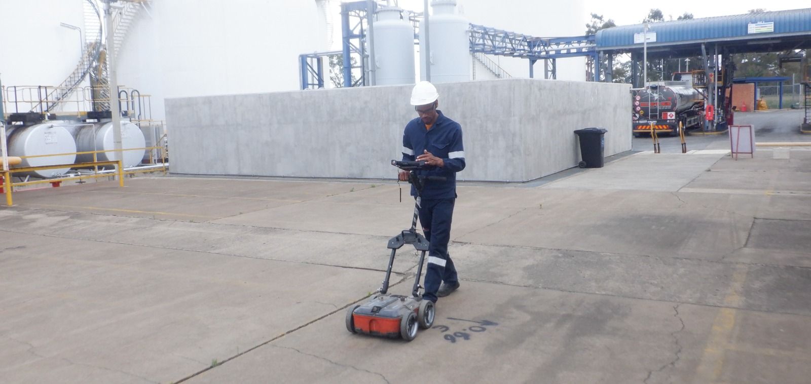 An HD GPR technician scanning a concrete yard with a ground penetrating radar cart, with storage tanks behind